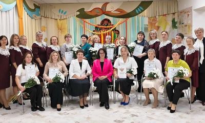 Групповое фото женщин с цветами и грамотами в празднично украшенном музыкальном зале детского сада.