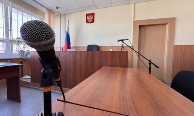Микрофон на стойке в пустом зале судебных заседаний, на стене герб России.